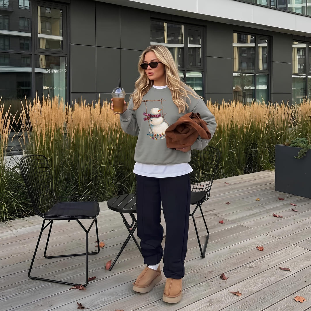 Woman wearing the Swing Season™ sweatshirt by Blossombless.co in Grey with dark jeans and sunglasses, holding a coffee and a brown bag on a wooden deck lined with tall grasses