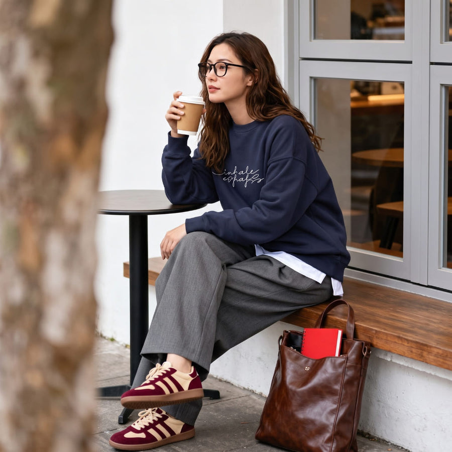 Woman wearing the Inhale Exhale™ oversized sweatshirt by Blossombless.co in Navy with gray trousers and burgundy sneakers, sitting at a café window with a coffee and a brown leather bag