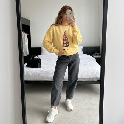 Woman wearing the Pieced Together™ sweatshirt by Blossombless.co in Butter with dark gray jeans and white sneakers, taking a mirror selfie in a bedroom