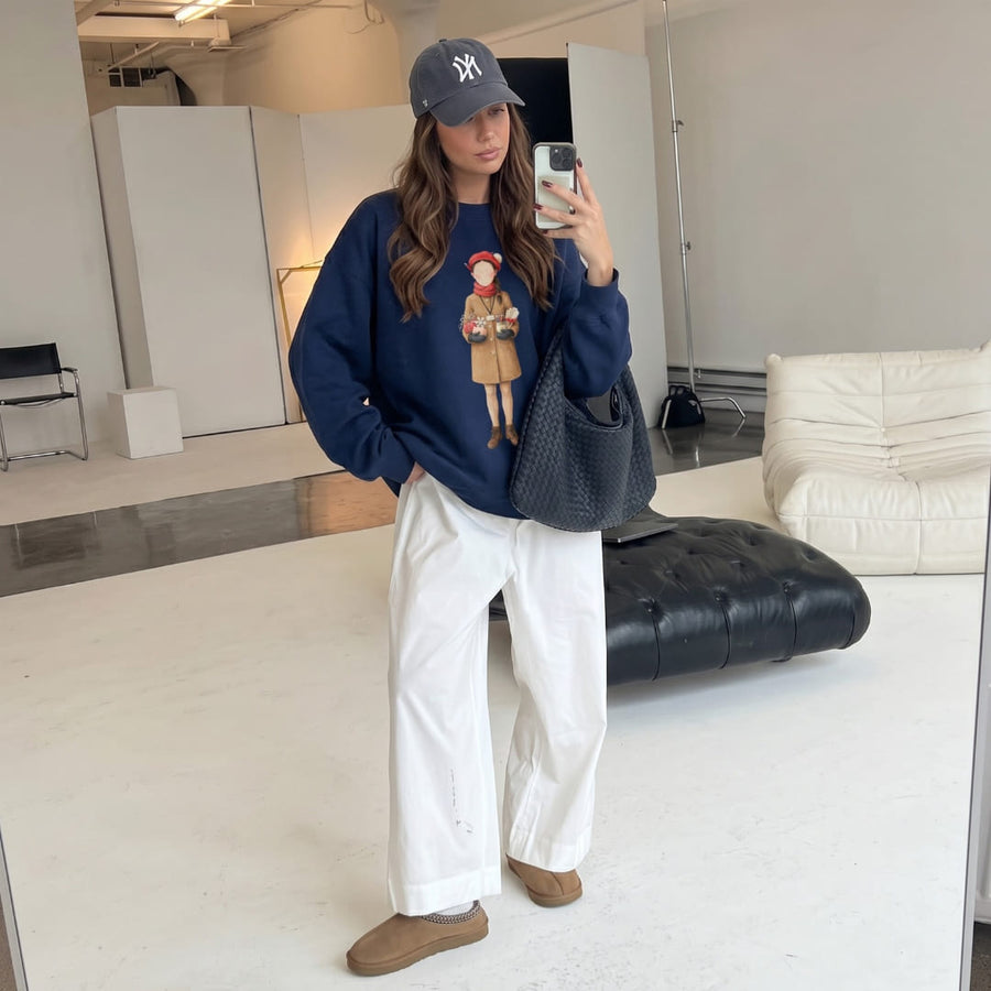 Woman wearing the Last Bell Before Break™ sweatshirt by Blossombless.co in True Navy with a navy cap, white wide-leg trousers and tan suede shoes, taking a mirror selfie in a bright minimalist room