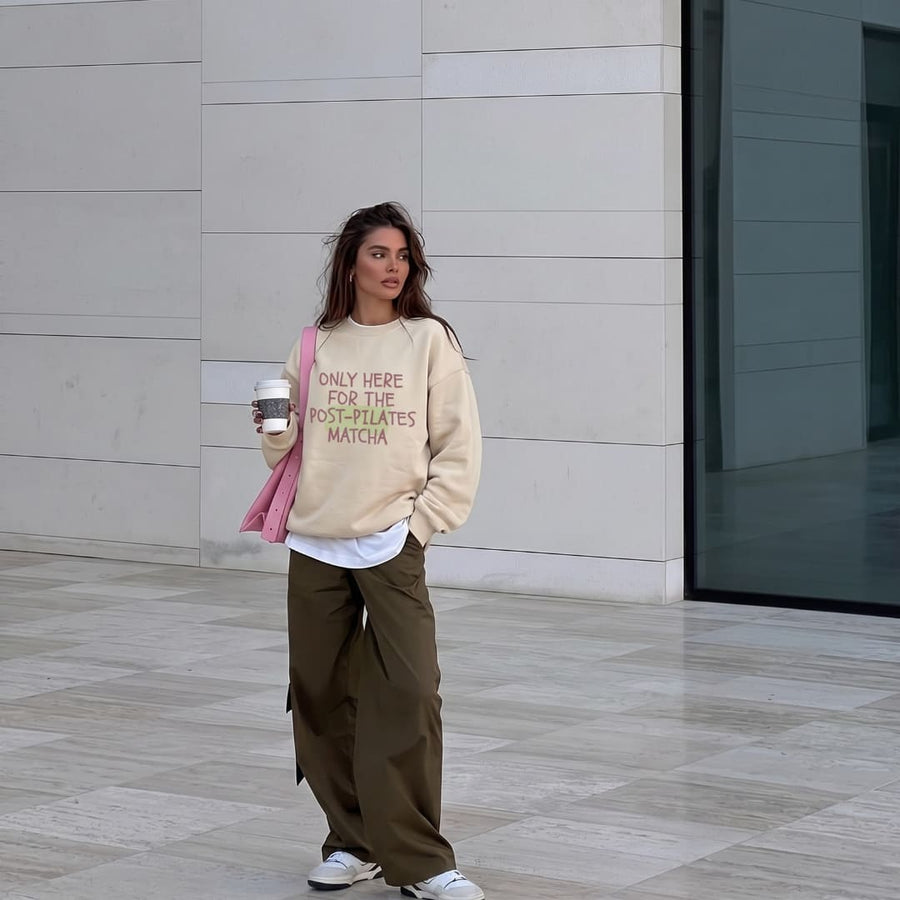 Woman wearing the Post-Pilates Matcha™ oversized sweatshirt by Blossombless.co in Dust with olive cargo pants and a pink shoulder bag, walking past a stone building with an iced drink