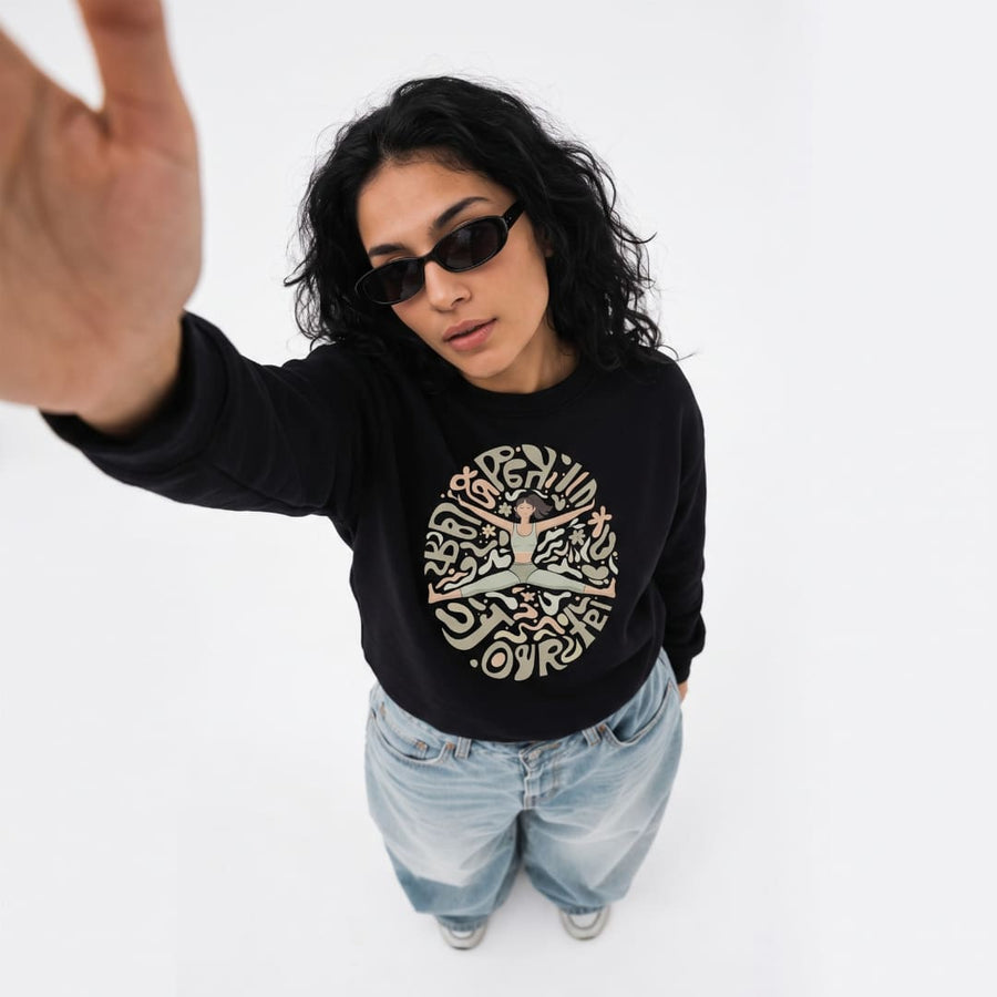 Woman wearing the Open In Yourself™ oversized sweatshirt by Blossombless.co in Vintage Black with sunglasses and baggy light-wash jeans, shot from above as she reaches toward the camera
