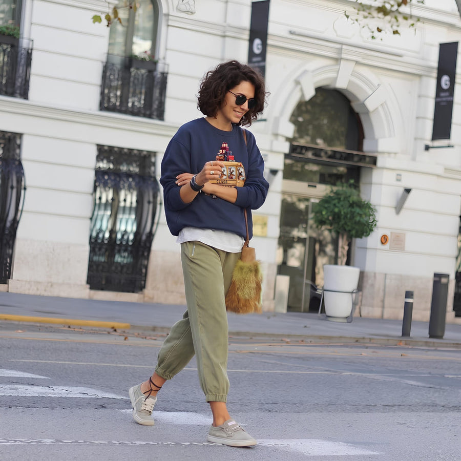 Woman wearing the Class Dismissed™ sweatshirt by Blossombless.co in True Navy with olive cargo pants and sneakers, walking across a city street lined with white stone buildings