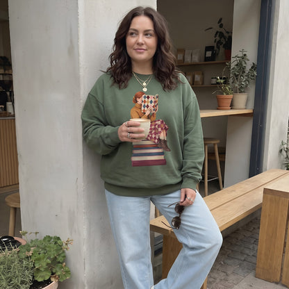 Woman wearing the Between Chapters™ sweatshirt by Blossombless.co in Blue Spruce with light-wash jeans, leaning against a wall outside a café