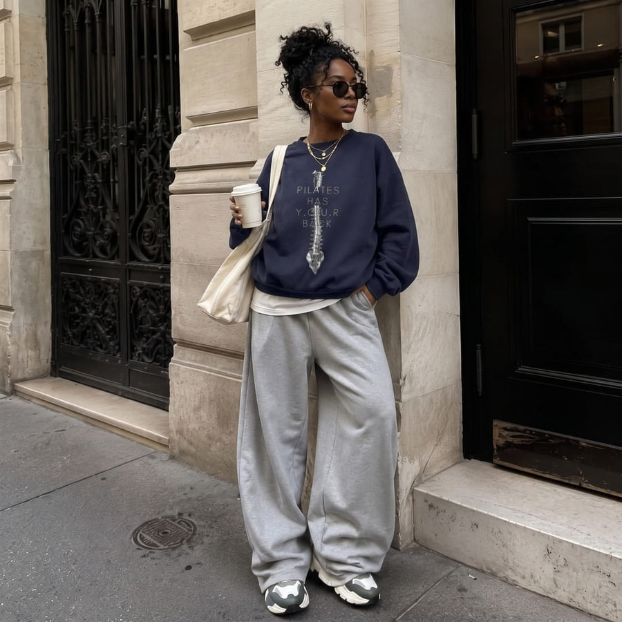 Woman wearing the Pilates Spine™ oversized sweatshirt by Blossombless.co in Navy with gray wide-leg sweatpants and sunglasses, holding a coffee outside a stone building