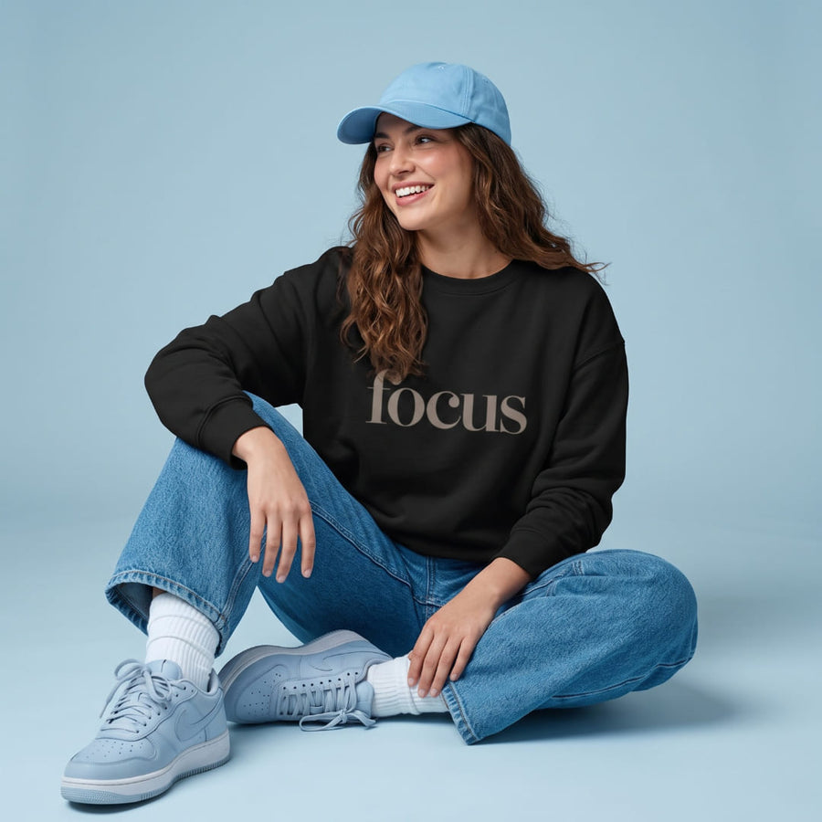 Focus™ oversized sweatshirt by Blossombless.co in Vintage Black, lifestyle: woman in jeans and a blue cap seated on a soft blue backdrop