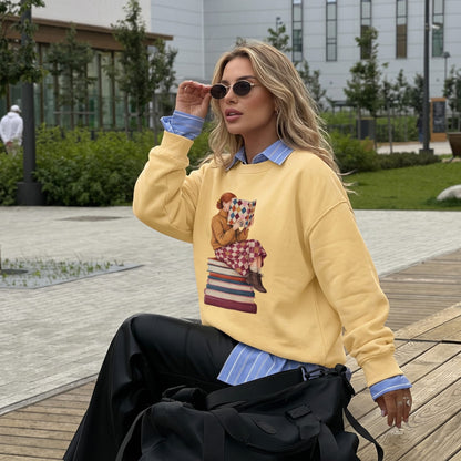 Woman wearing the Between Chapters™ sweatshirt by Blossombless.co in Butter over a blue button-down with black trousers and sunglasses, sitting outdoors