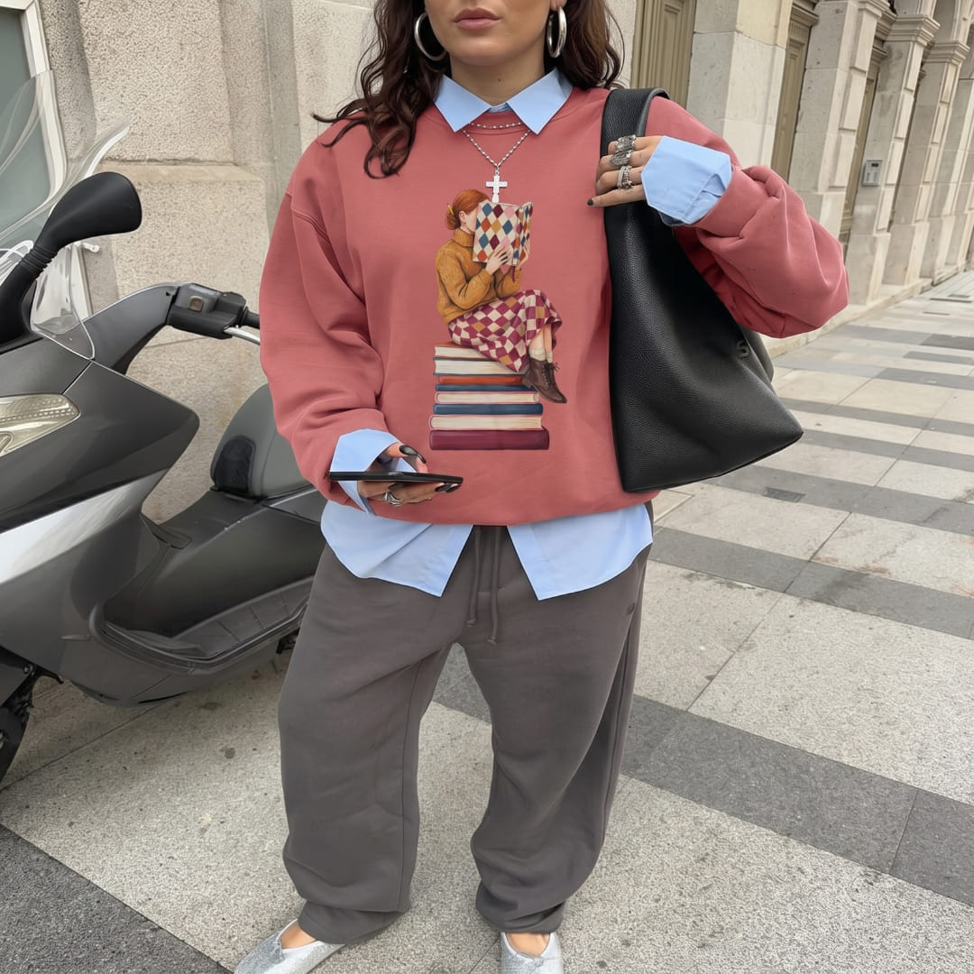Woman wearing the Between Chapters™ sweatshirt by Blossombless.co in Crimson over a blue button-down with brown trousers and a black bag, standing on a city street