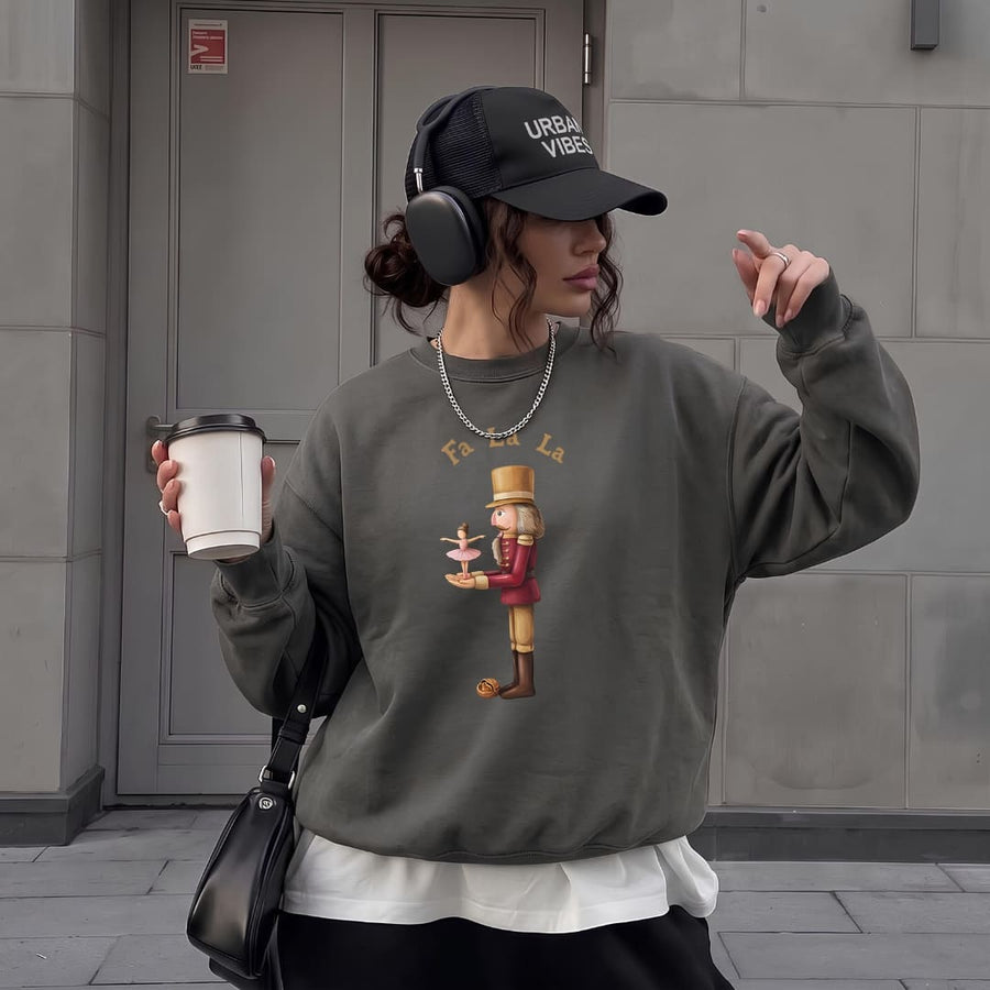 Woman in a black cap and headphones holding a coffee on a city sidewalk, wearing the Pas de Deux™ Nutcracker sweatshirt in Pepper
