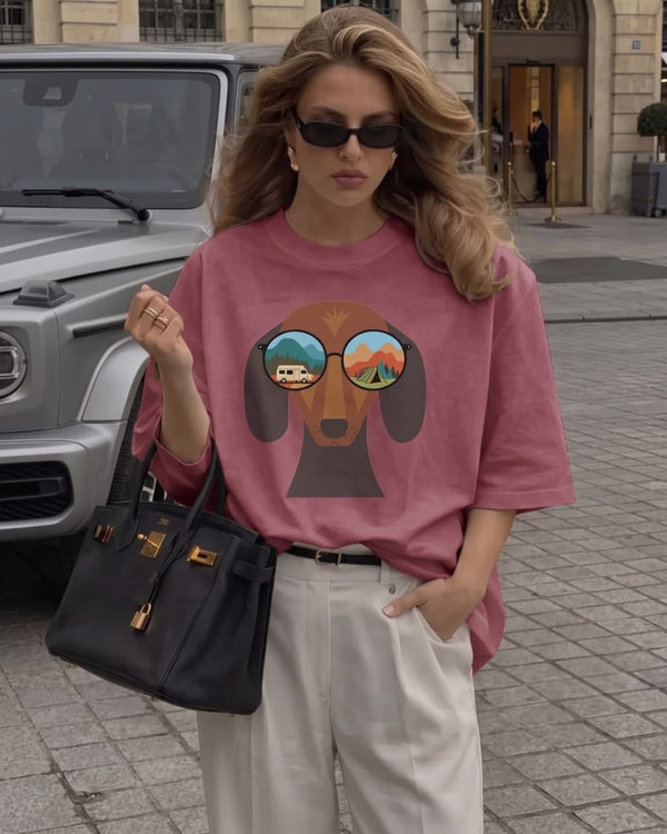 A woman with a blonde blowout and sunglasses wears The Adventure Hound™ tee in berry tucked into cream trousers, holding a black leather handbag on a cobblestone square.