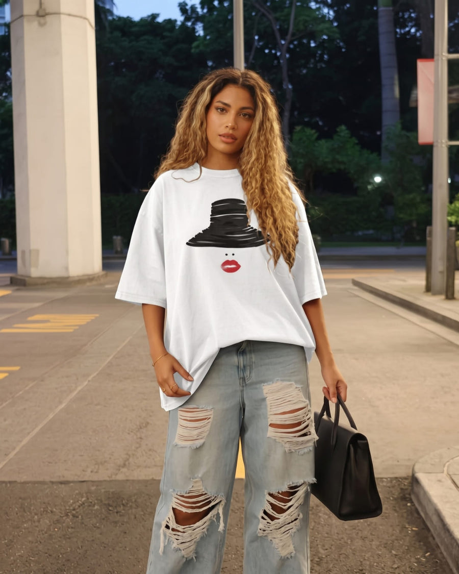 A woman with long curly blonde hair wears an oversized white The French Riviera™ tee with ripped light-wash jeans and a black handbag on a city street at dusk.