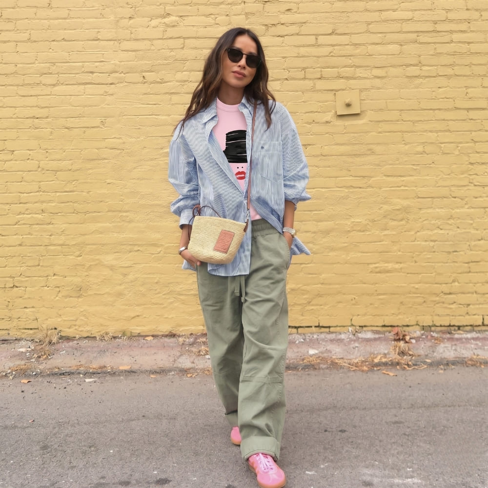 Woman standing against a yellow brick wall wearing a denim jacket, pink shirt, green pants, and pink shoes.
