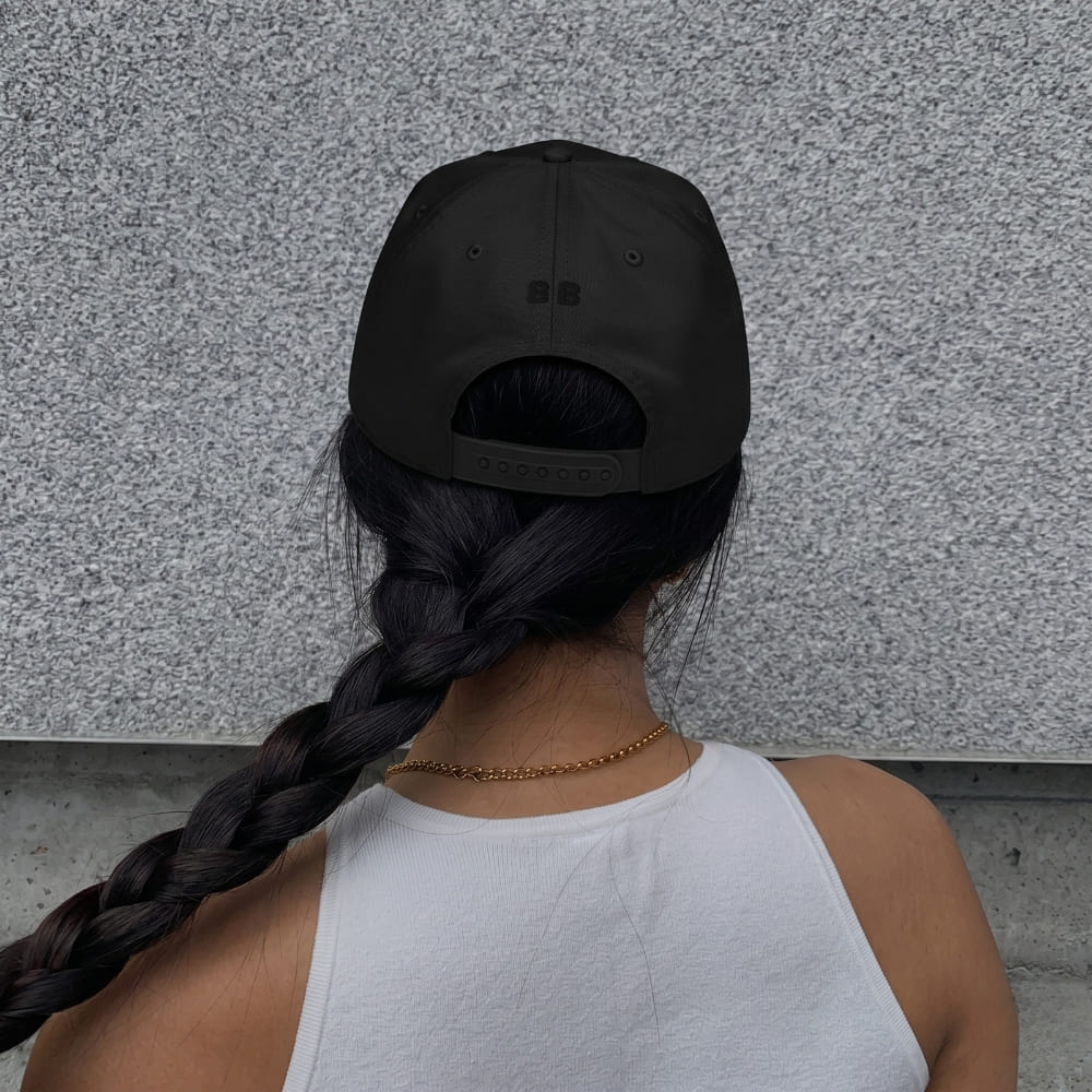 Woman wearing a black cap and white tank top with braided hair, standing against a gray wall.