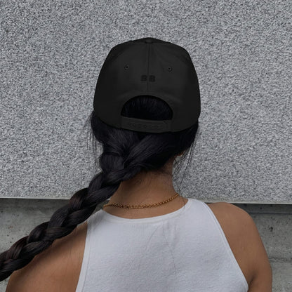 Woman wearing a black cap and white tank top with braided hair, standing against a gray wall.