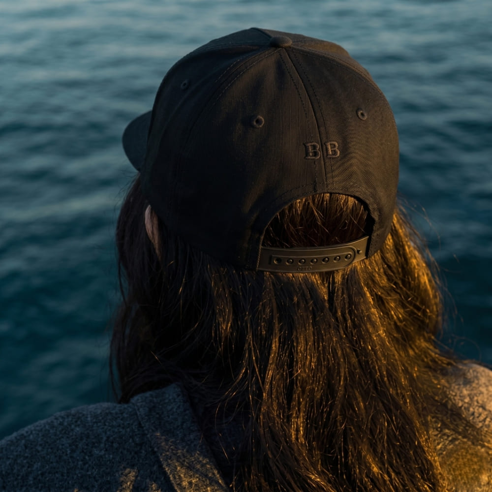 Woman wearing a black cap with 'BB' logo, standing by water