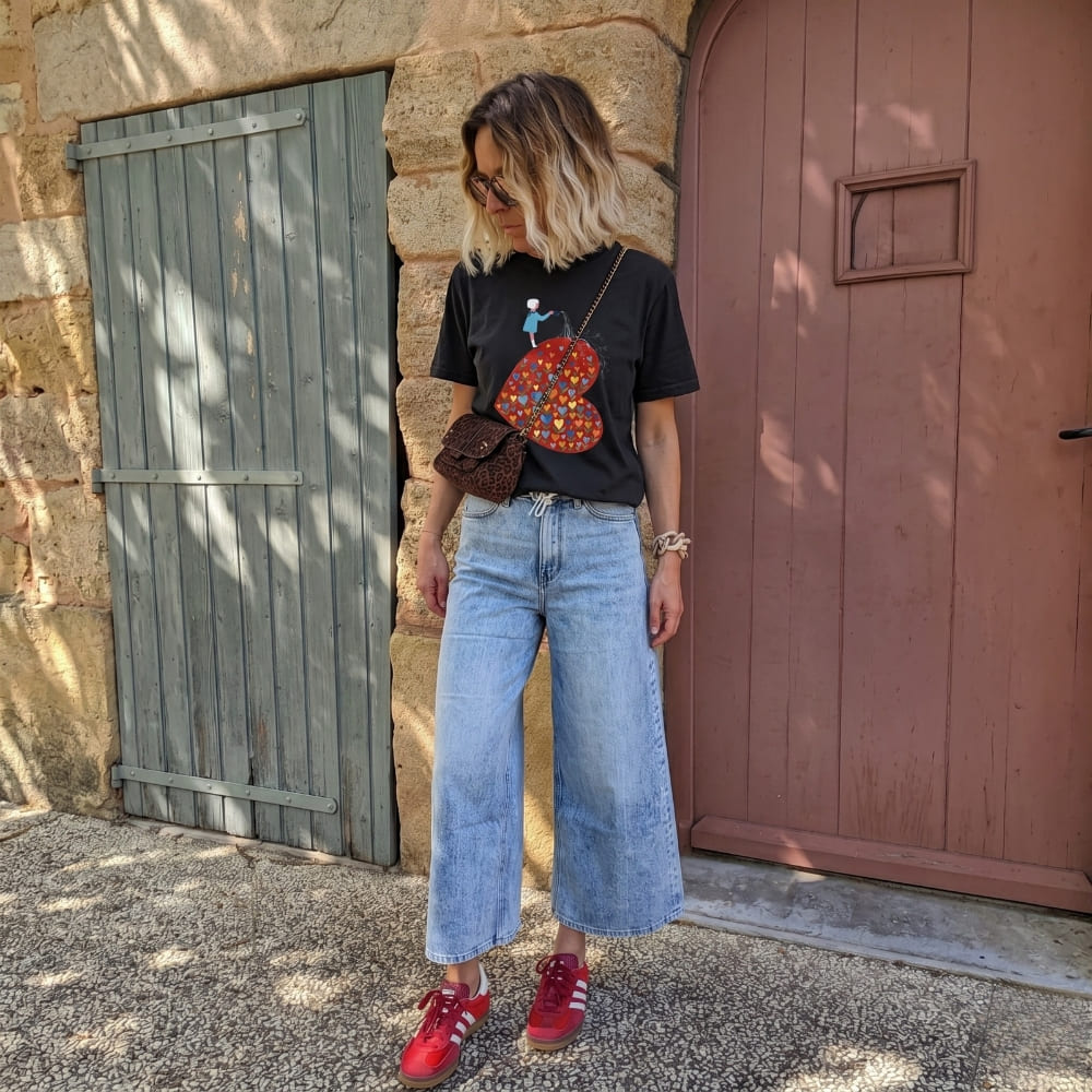 Person wearing a black t-shirt with a colorful heart design, blue jeans, and red shoes standing in front of a stone wall.