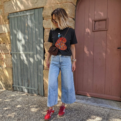 Person wearing a black t-shirt with a colorful heart design, blue jeans, and red shoes standing in front of a stone wall.