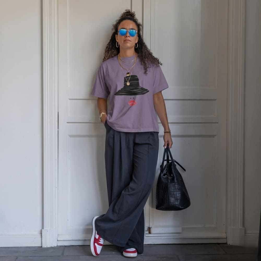 Woman wearing a purple t-shirt with a graphic design, dark pants, and red sneakers, holding a black handbag against a white wall.