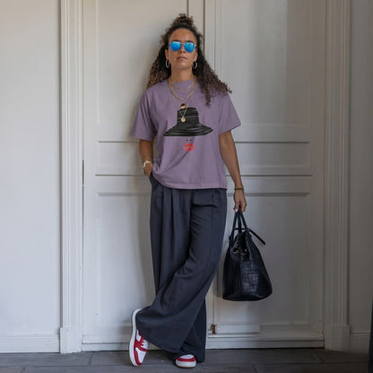 Woman wearing a purple t-shirt with a graphic design, dark pants, and red sneakers, holding a black handbag against a white wall.