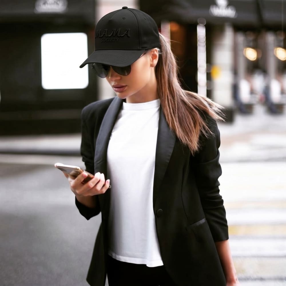 Woman wearing a black cap and black blazer, looking at her phone in an urban setting.