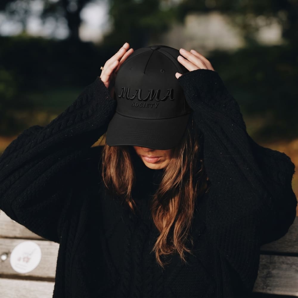 Woman wearing a black cap with 'Mama Society' text, standing outdoors.