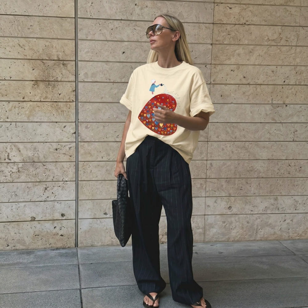 Woman wearing a beige t-shirt with a colorful heart design and dark pants against a stone wall.
