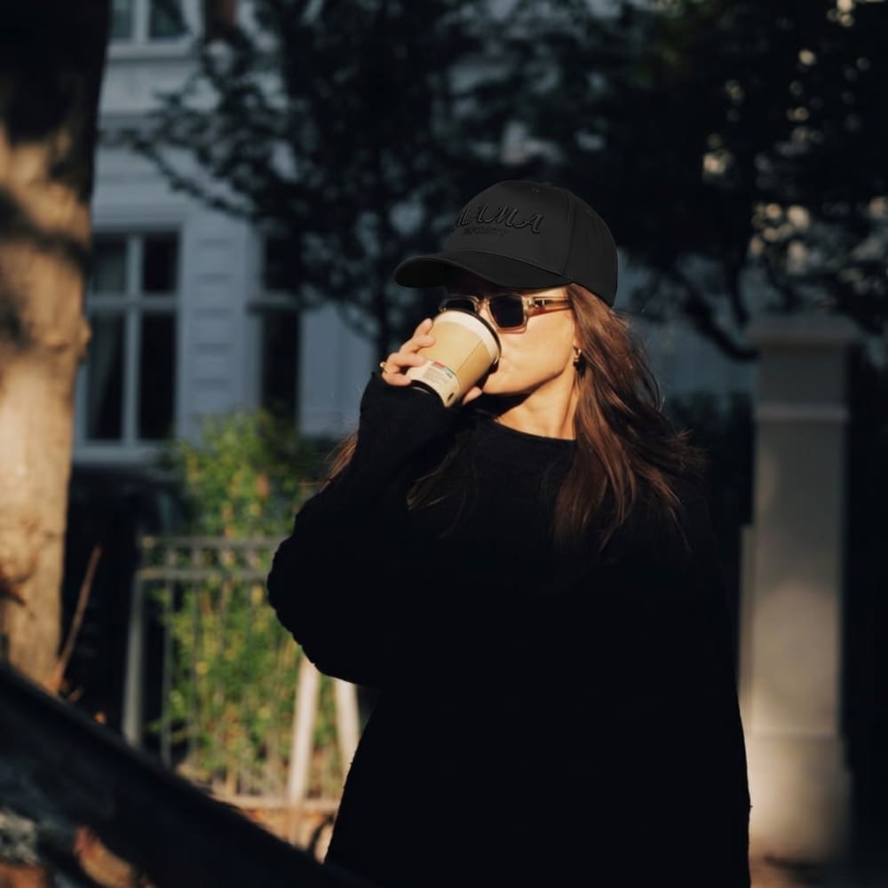 Person wearing a black cap and sunglasses, drinking from a coffee cup outdoors.