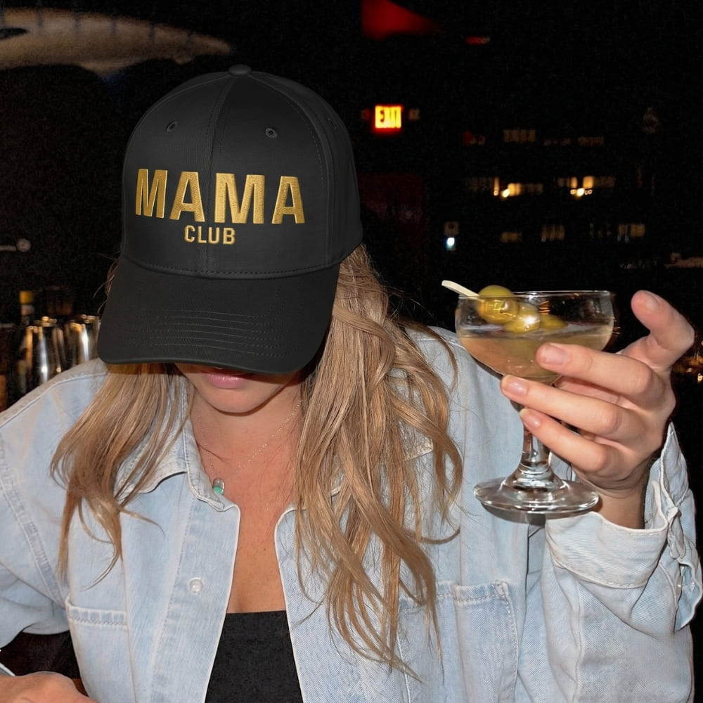 Person wearing a 'MAMA CLUB' cap holding a cocktail at night.