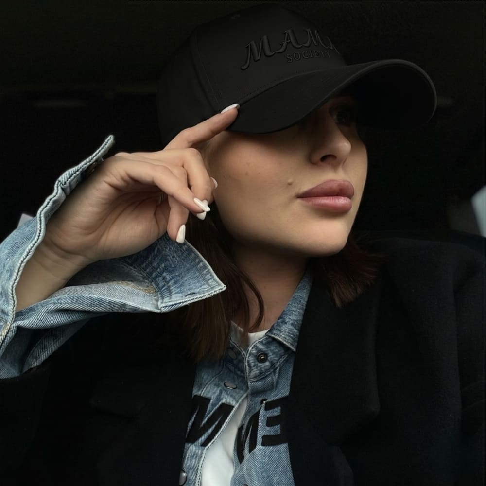 Woman wearing a black cap with 'MAMA SOCIETY' branding, posing against a dark background.