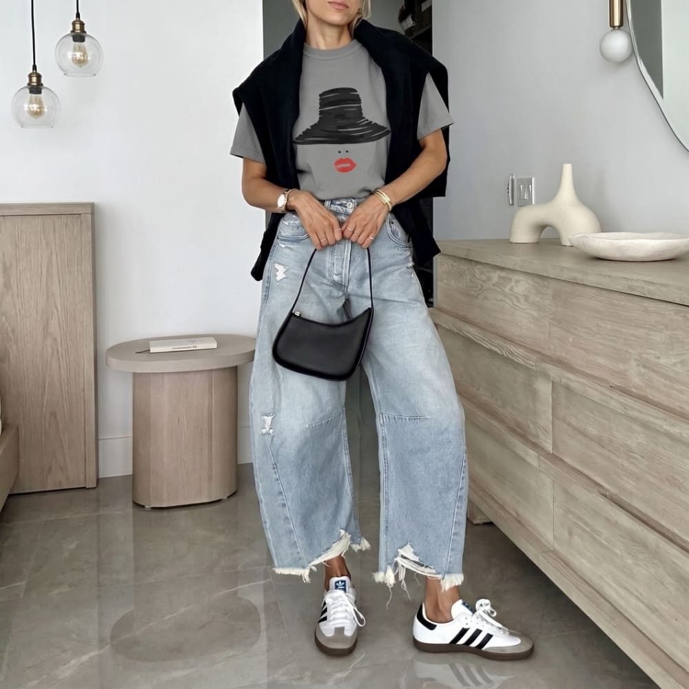 Woman wearing a graphic t-shirt, black jacket, light blue jeans, and white sneakers in a room with wooden furniture.