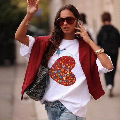 Woman talking on a phone, wearing a white t-shirt with a colorful heart design, red jacket, and sunglasses.