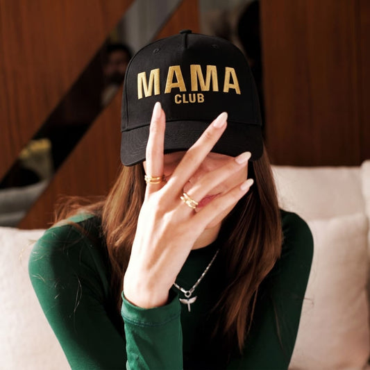 Person wearing a black cap with 'MAMA CLUB' text, holding hand to face, blurred background