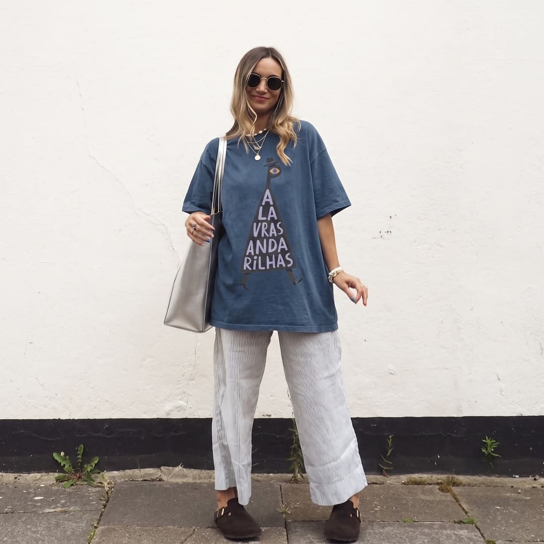 A woman stands outdoors against a white wall wearing an oversized blue graphic T-shirt with white text, light gray wide-leg pants, dark brown loafers, sunglasses, layered necklaces, and a silver shoulder bag.