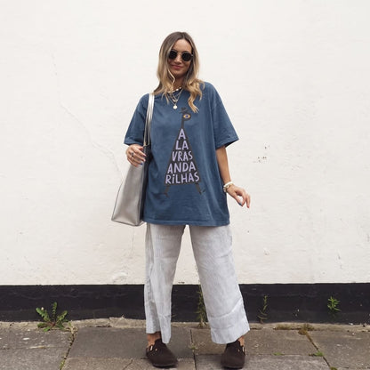 A woman stands outdoors against a white wall wearing an oversized blue graphic T-shirt with white text, light gray wide-leg pants, dark brown loafers, sunglasses, layered necklaces, and a silver shoulder bag.