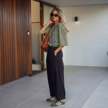 A woman stands in a modern outdoor walkway wearing an olive green graphic t-shirt, black wide-leg pants, olive green sneakers, and carrying a brown leather shoulder bag.