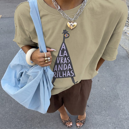 A person wearing an oversized beige graphic T-shirt with a black triangular design and the text 'A LA VRAS ANDA RILHAS', paired with brown shorts, a light blue tote bag, layered silver necklaces, and leopard-print flat sandals on a gray pavement background.