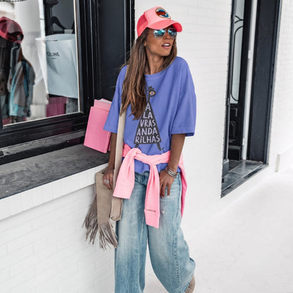 A woman wearing an oversized purple graphic T-shirt with a pink shirt tied around her waist, blue jeans, a coral pink cap, and sunglasses, standing in front of a white brick wall.