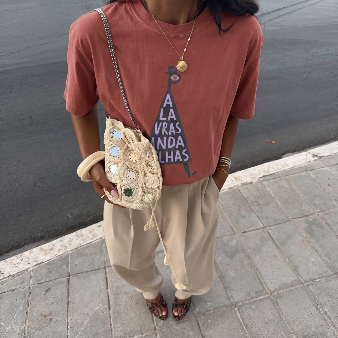 A person standing outdoors on a paved sidewalk next to a road, wearing an oversized dusty rose graphic T-shirt, loose beige pants, and leopard-print flats, while carrying a small embellished cream bucket bag.