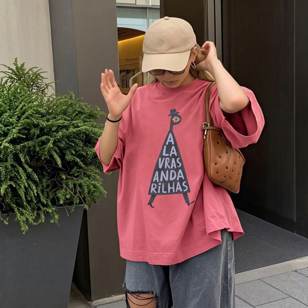A woman wearing an oversized pink graphic t-shirt with a black triangular text design, paired with a beige baseball cap, ripped gray jeans, and a brown leather shoulder bag, standing outdoors.