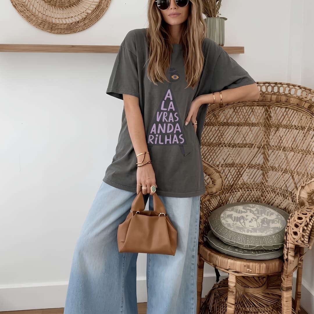 A woman wearing a dark gray oversized graphic t-shirt with purple text, light blue wide-leg jeans, and sunglasses, holding a small brown leather handbag. She is posed next to a woven rattan chair in a bright room.
