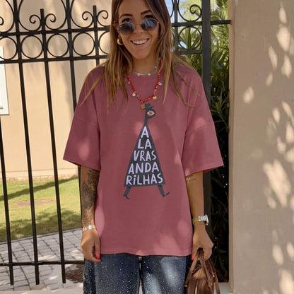 A woman wearing an oversized dusty rose t-shirt with a graphic of a black triangle containing the Portuguese text 'A LA VRAS ANDA RILHAS' in white, paired with blue jeans and sunglasses, standing outdoors in front of a black wrought-iron gate.