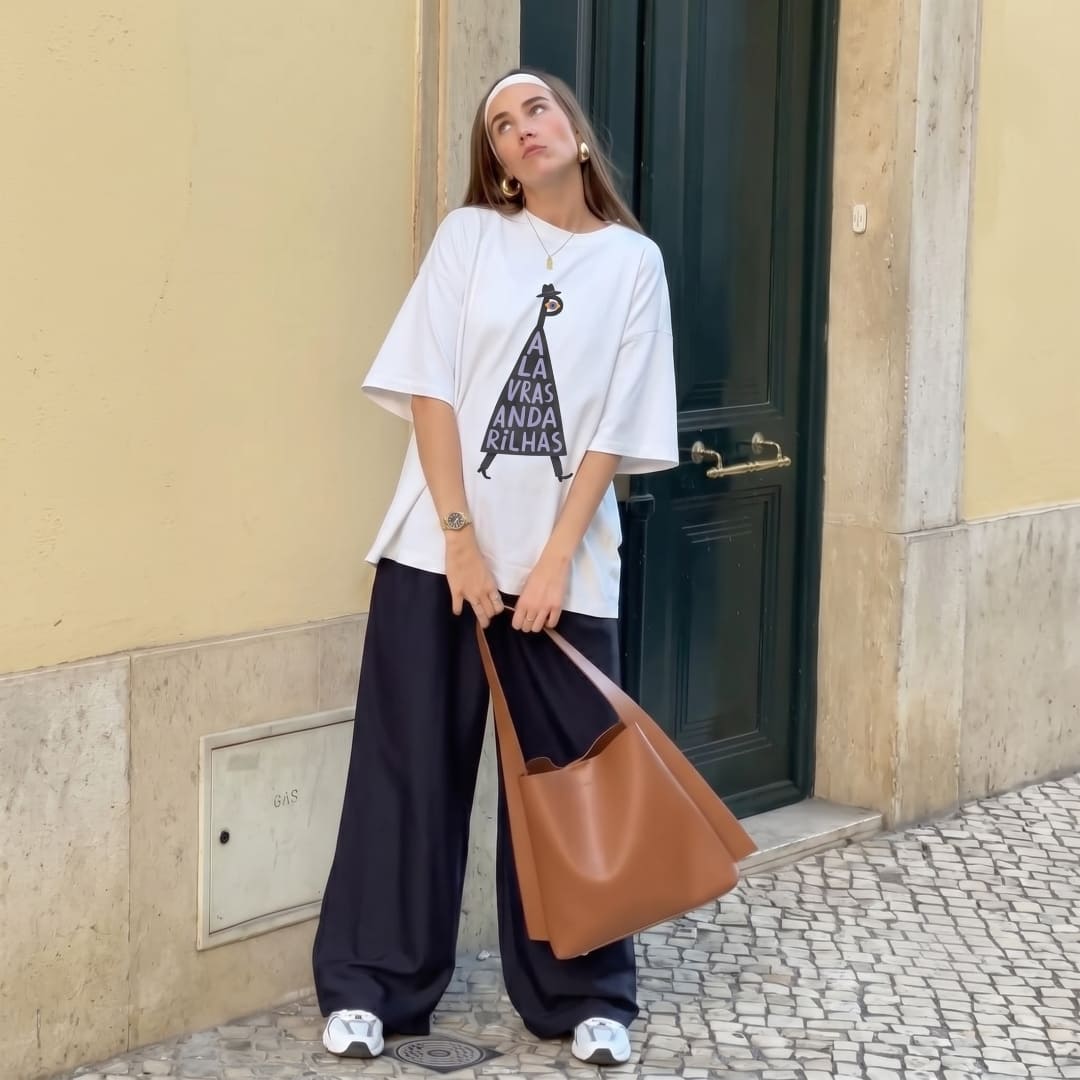 A woman stands on a cobblestone street in front of a beige wall and dark green door, wearing an oversized white graphic T-shirt, loose dark wide-leg pants, white sneakers, and carrying a large tan tote bag.