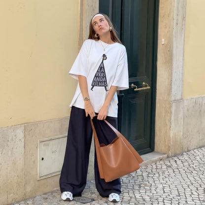 A woman stands on a cobblestone street in front of a beige wall and dark green door, wearing an oversized white graphic T-shirt, loose dark wide-leg pants, white sneakers, and carrying a large tan tote bag.