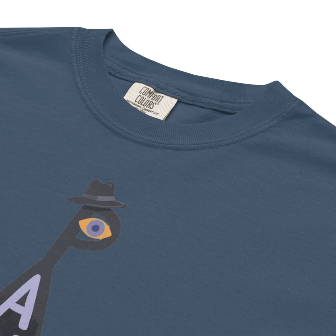 Close-up of a slate blue crewneck t-shirt featuring a graphic of a stylized figure with a black fedora and an eye graphic on the chest. The inside collar shows a brand label for 'Combat Colors'.