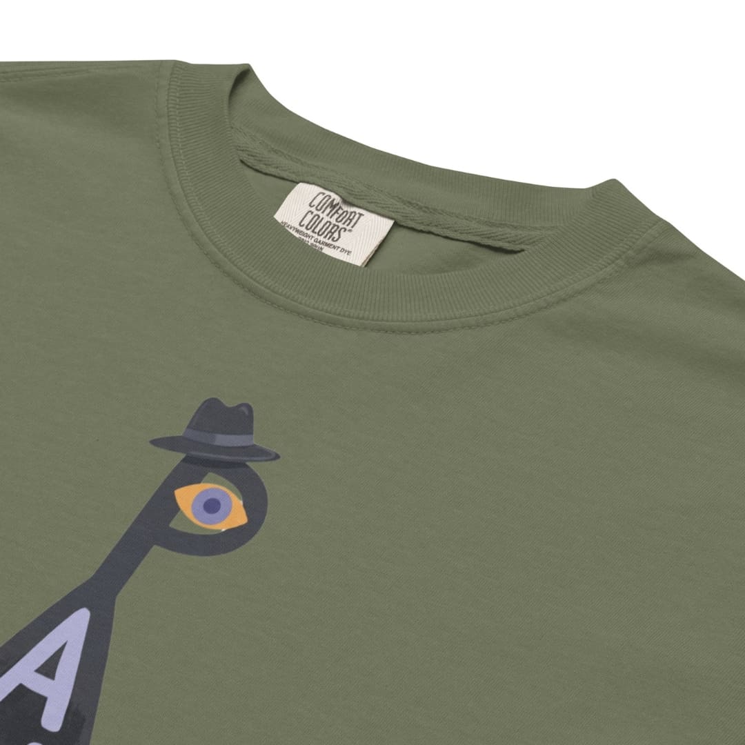 Close-up of an olive green t-shirt featuring a graphic of a bird wearing a black fedora hat and a single eye, with a visible Combat Colors brand label inside the collar.
