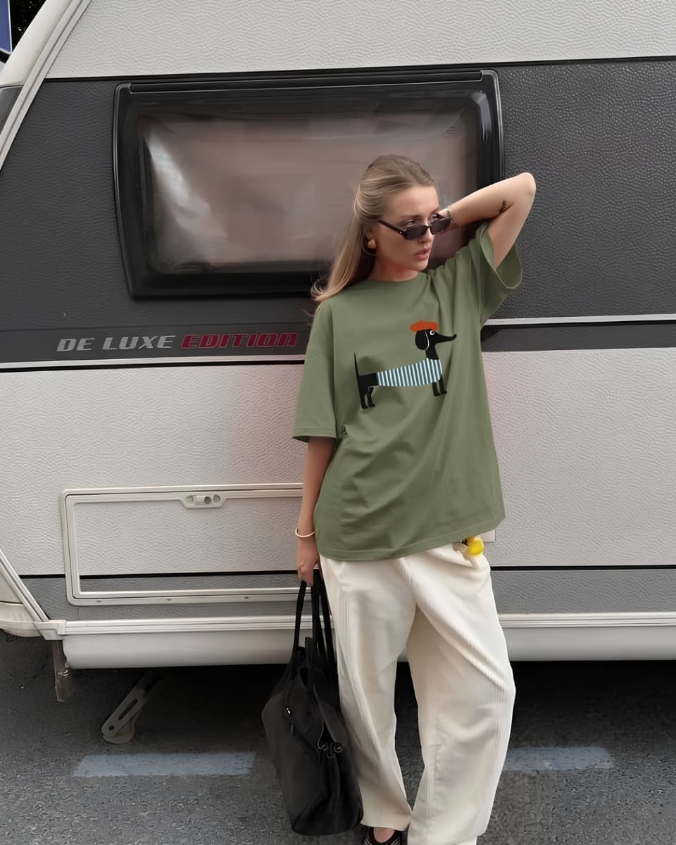 Woman in an olive graphic tee, cream wide-leg pants and sunglasses standing beside a vintage caravan