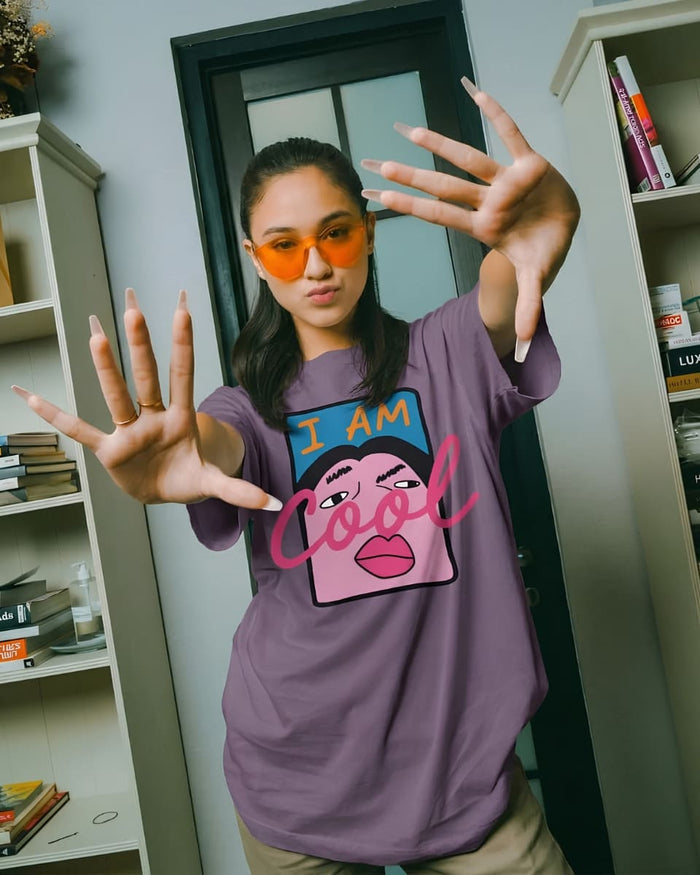 Model wearing an oversized purple graphic tee with orange sunglasses, hand raised toward camera