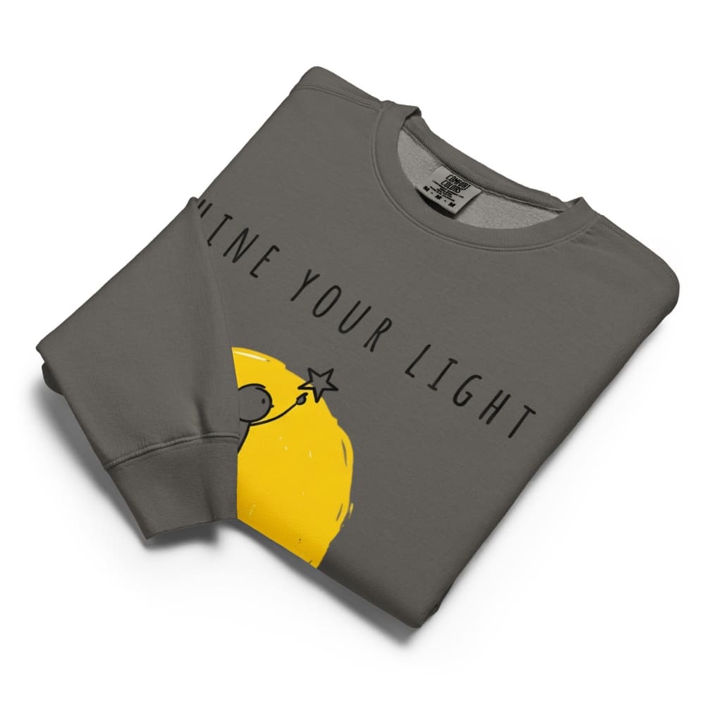 Folded gray sweatshirt with yellow graphic and text 'Shine Your Light' on a white background