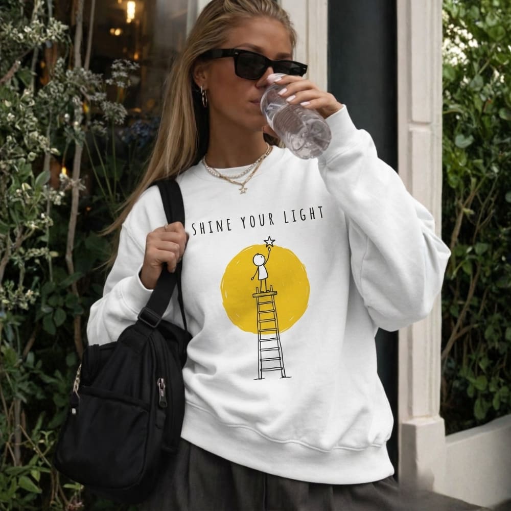 Woman wearing a white sweatshirt with a graphic design and text, drinking from a water bottle outdoors.
