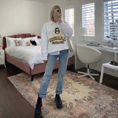Person wearing a white sweatshirt with graphic design and ripped jeans in a bedroom.